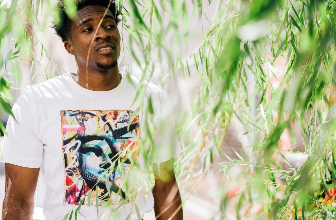 Man wearing a white abstract art graphic T-shirt among green foliage, suitable for colorful Print on Demand Streetwear and artist-led apparel collections