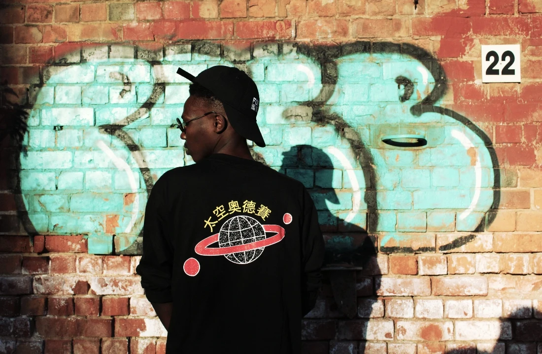 Man wearing a black long-sleeve shirt with a large space-inspired back graphic beside a graffiti-covered brick wall