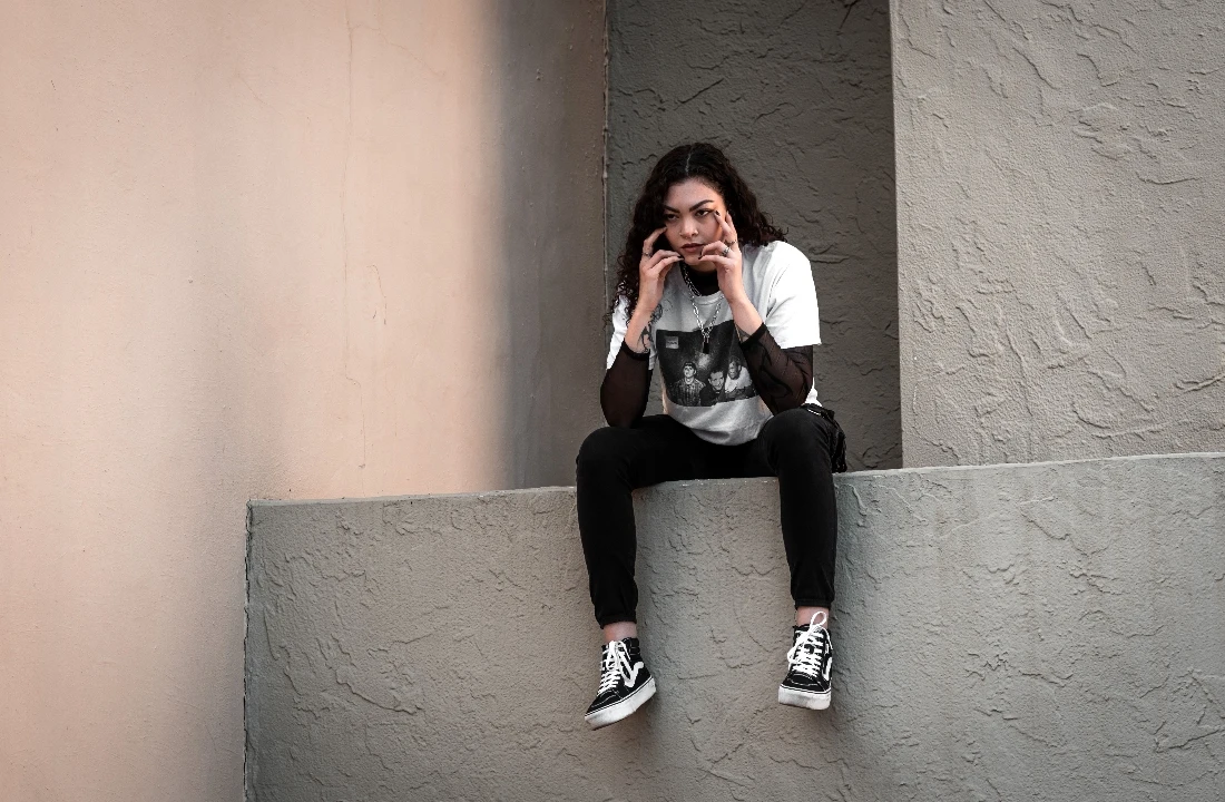 Woman wearing a layered white graphic T-shirt and black streetwear outfit while seated against a modern concrete wall