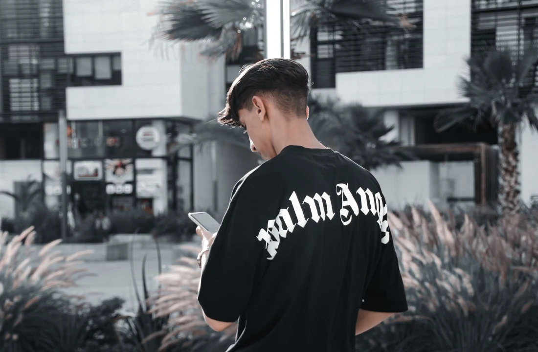 Young man checking his phone while wearing a black oversized T-shirt with large gothic lettering across the back