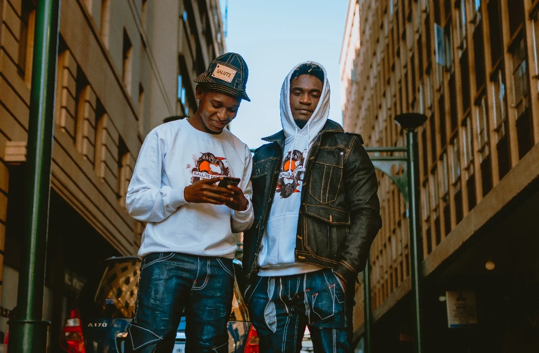 Two men wearing coordinated graphic streetwear, including a white hoodie layered under a dark denim jacket