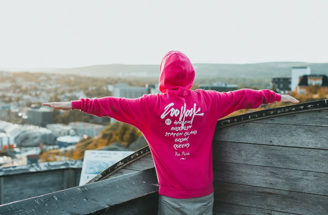Person wearing a bright pink printed hoodie with arms extended above an urban city view