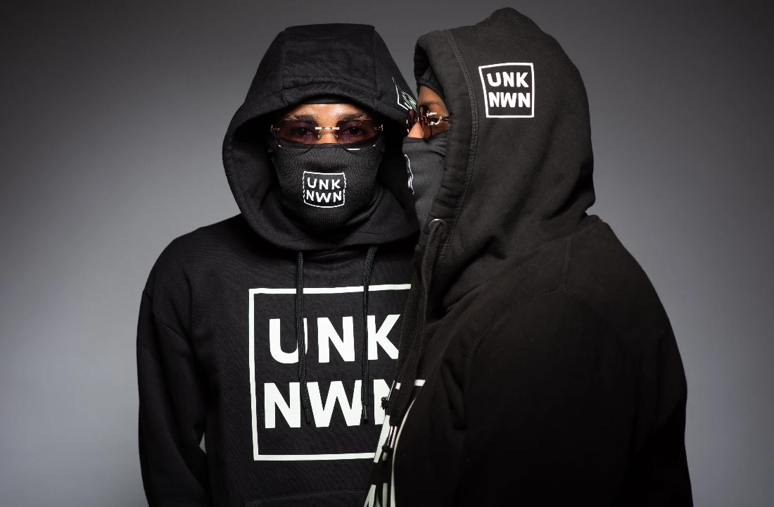 Two models wearing coordinated black branded hoodies and matching face coverings in a studio setting