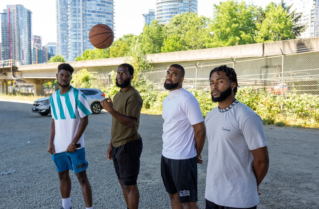 Four men wearing casual athletic tops and streetwear shorts outdoors with a basketball in the air
