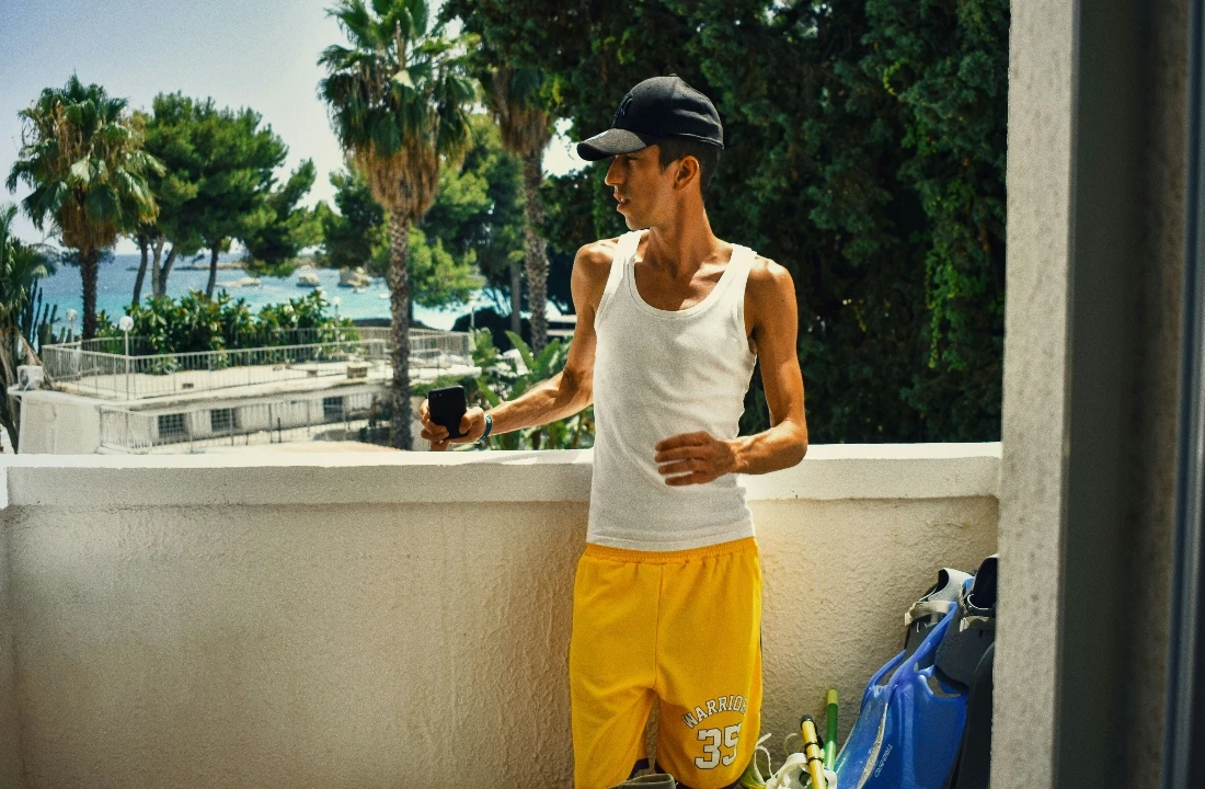 Man wearing bright yellow streetwear shorts and a white tank top on a sunny balcony near the coast