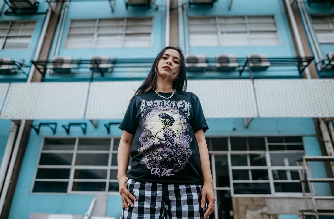 Woman wearing a black POD Streetwear graphic T-shirt and checkered pants, standing in front of a blue urban building with a bold streetwear look