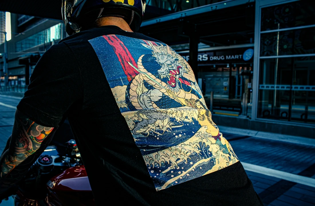 Close-up back view of a rider wearing a black Streetwear Print on Demand T-shirt with a large dragon print while sitting on a motorcycle