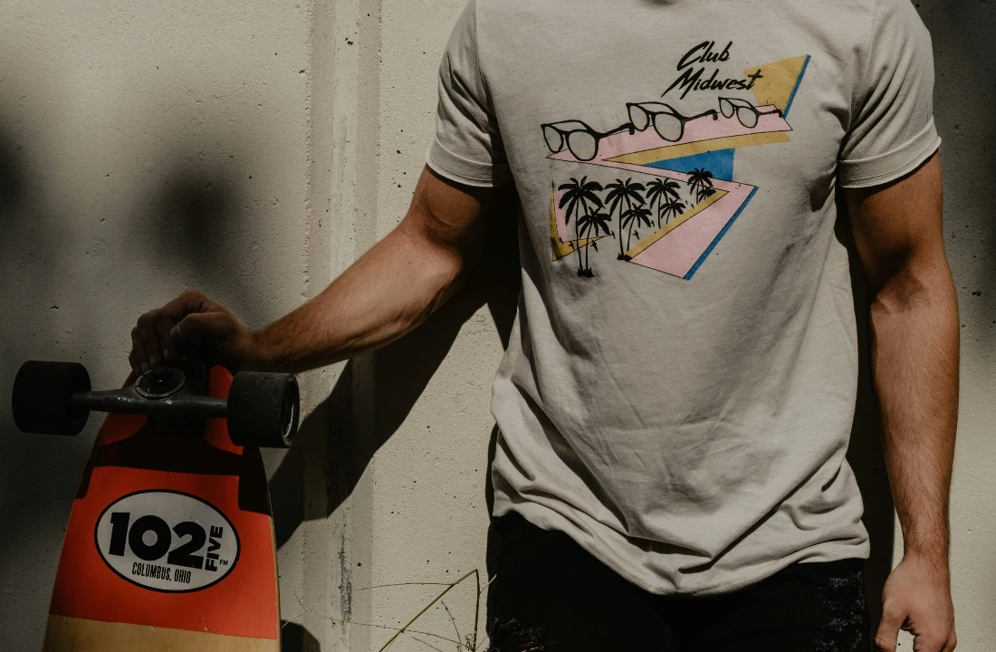 A man standing with a skateboard wearing a beige Print on Demand Streetwear T-shirt with a retro palm tree graphic