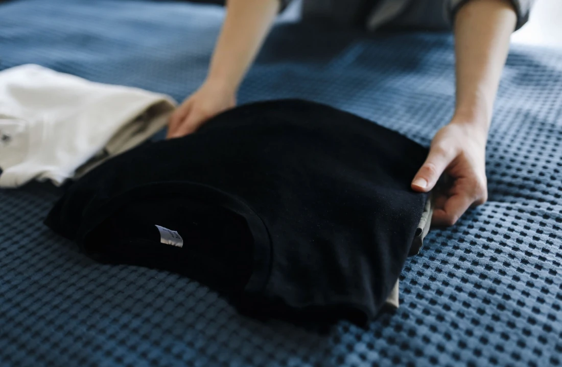 Hands folding a black Organic Cotton Custom T Shirts piece on a bed, showing a clean and minimal product presentation