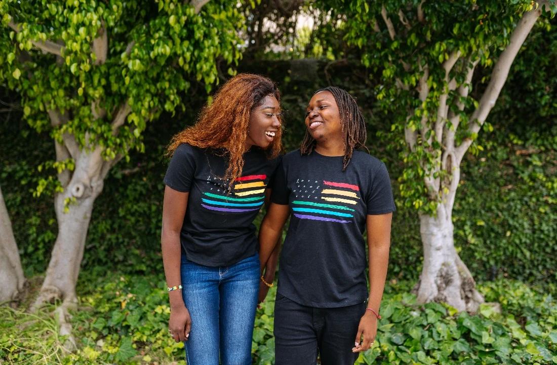 Two people wearing matching black Pride Apparel T shirts with rainbow stripe graphics outdoors