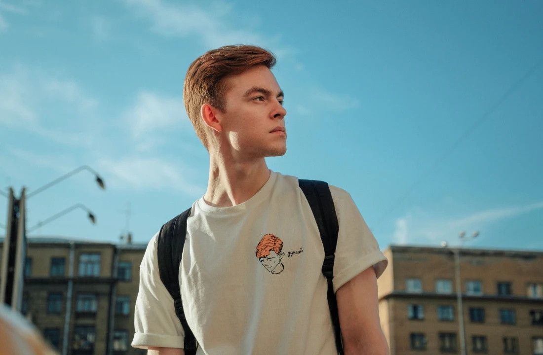 Young man wearing a light-colored print on demand streetwear T shirt with a small chest graphic and backpack in an urban setting
