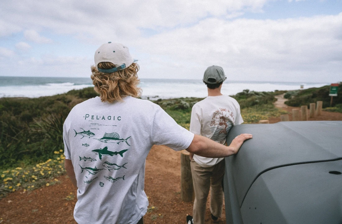 Two people wearing white POD T shirts with back graphic prints near a coastal landscape