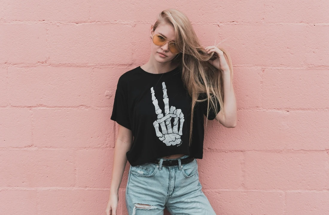 Woman wearing Custom Graphic Tees with a skeleton hand peace sign print against a pink wall