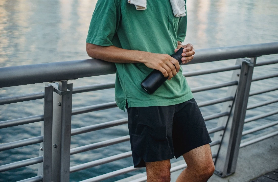 Man wearing black Fleece Shorts with utility-style pockets and a green oversized T-shirt beside the waterfront