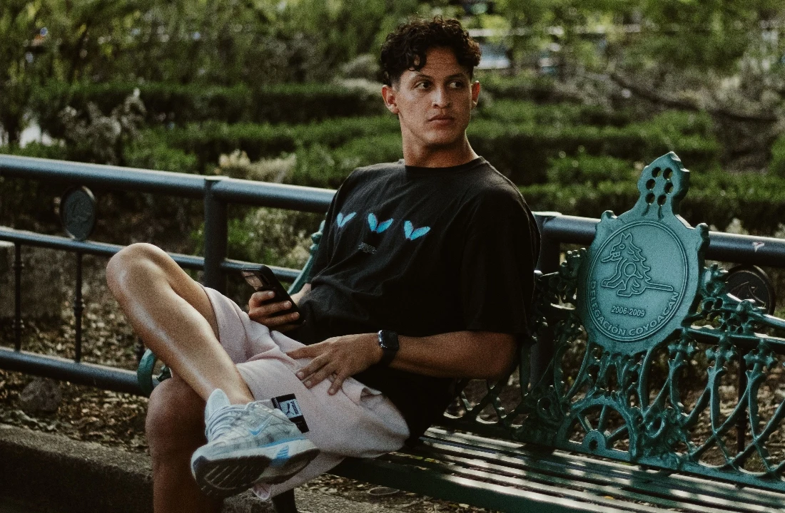 Man sitting on an outdoor bench wearing light neutral Fleece Shorts with a black graphic T-shirt